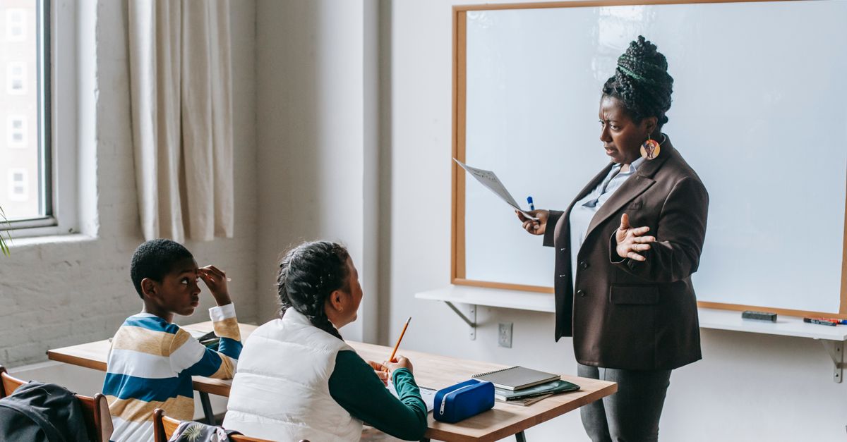 Adult African American female teacher in formal suit standing near whiteboard and explaining new material to multiethnic elementary pupils in light classroom