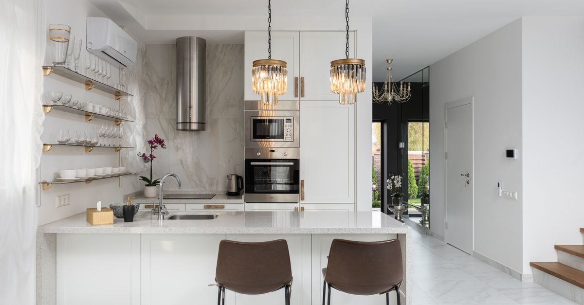 Interior of contemporary kitchen with modern appliances and chairs at white counter near wall with shelves in spacious apartment near stairs