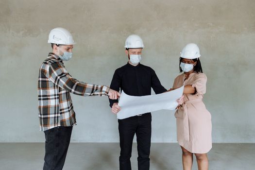 Three architects wear masks and helmets while reviewing construction plans indoors.