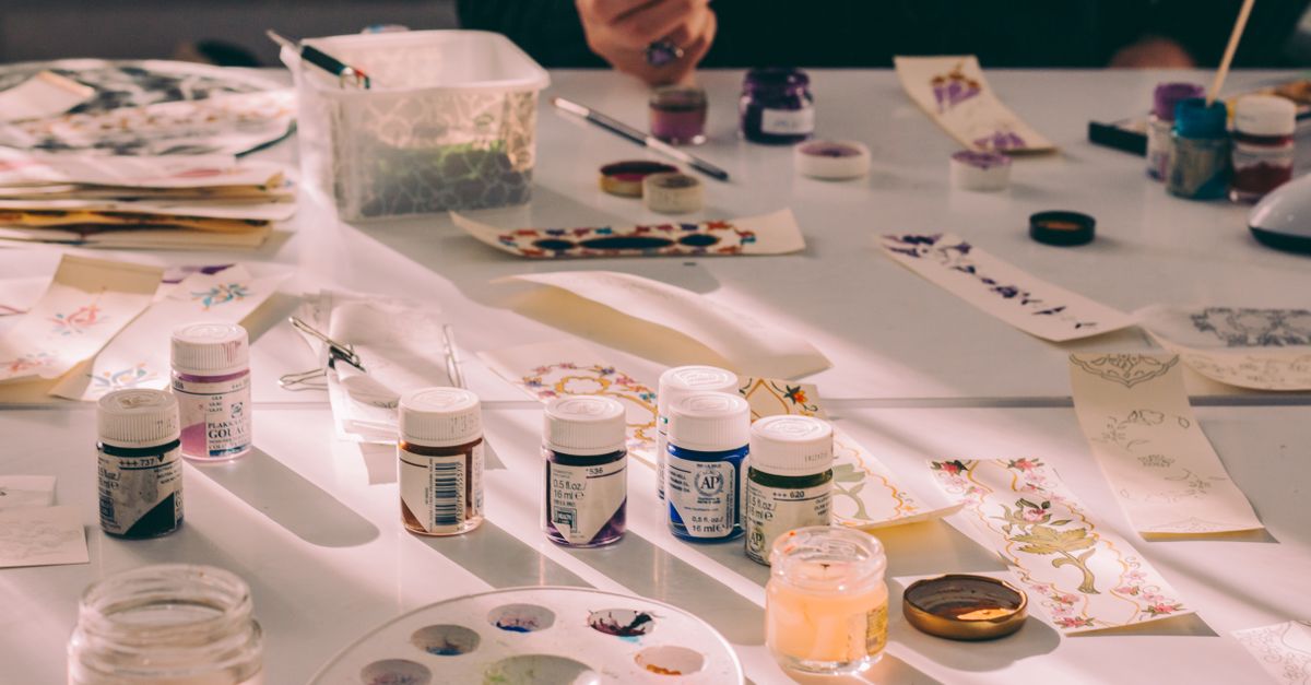 Art studio table with various paints, paper, and an artist's hand crafting designs.