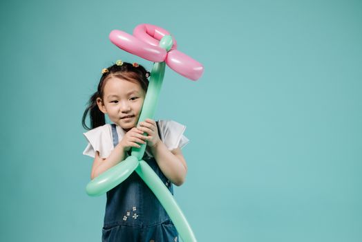 Adorable Asian girl in denim holding a pink and green balloon shaped like a flower against a teal backdrop.
