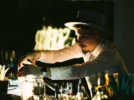 A skilled bartender wearing a hat mixes drinks behind the bar in a dimly lit club setting.