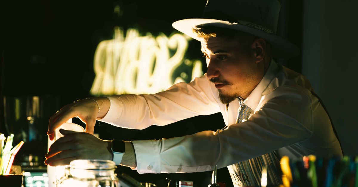 A skilled bartender wearing a hat mixes drinks behind the bar in a dimly lit club setting.
