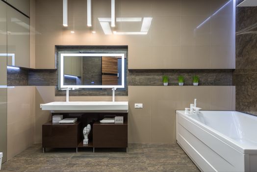 White bathtub placed near cabinet with sink at wall with mirror in light stylish bathroom with glowing illumination in apartment
