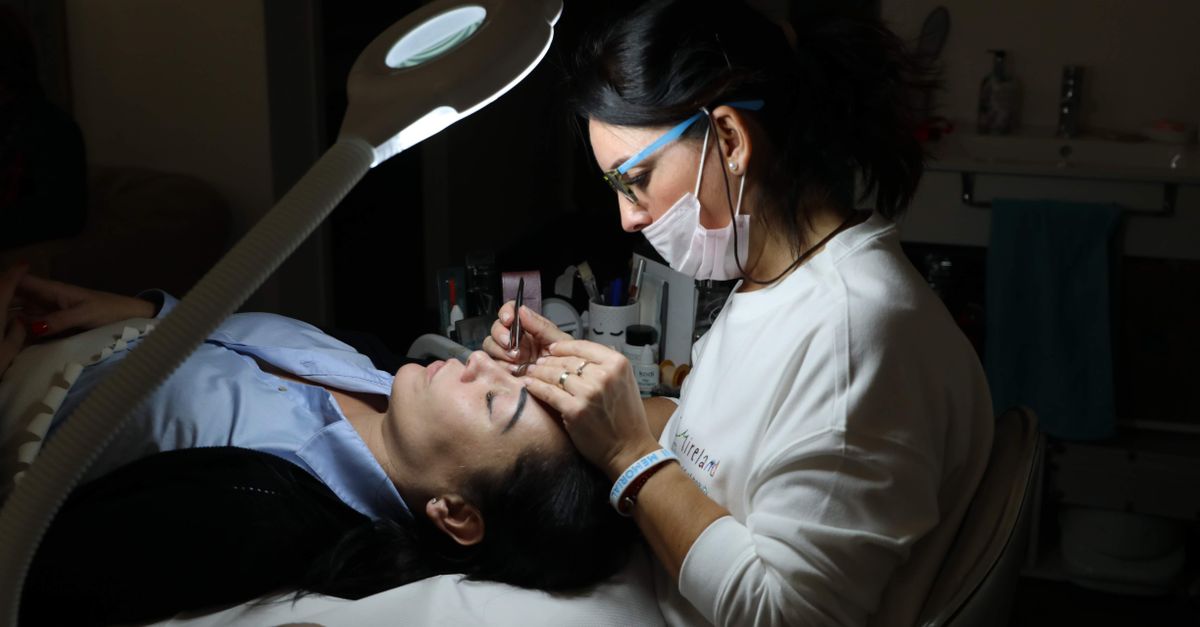 A beautician provides professional eyebrow care to a client in a salon, under focused lighting.