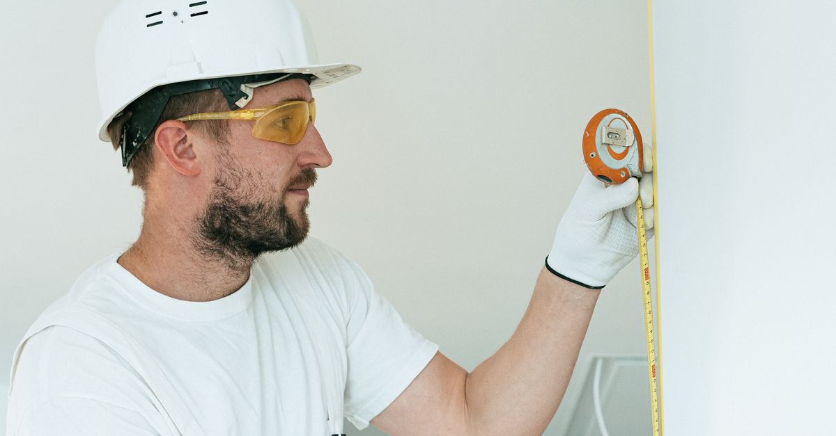 Construction worker with hard hat and goggles measuring a wall with a tape measure.