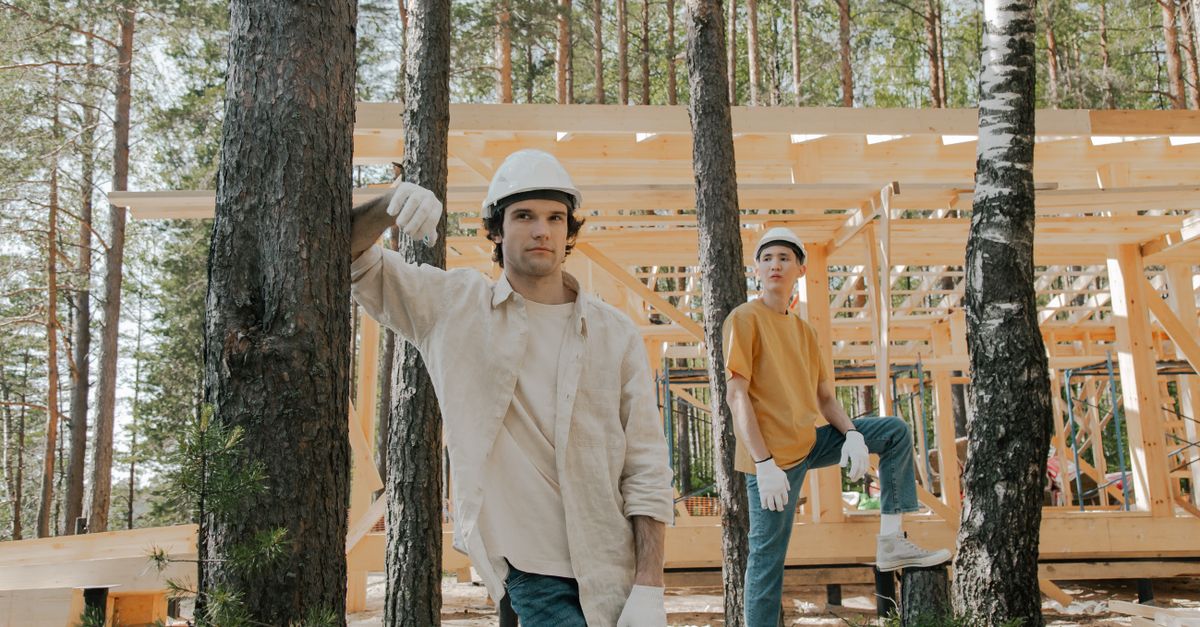 Two construction workers in protective gear at a forest site with wooden structure.
