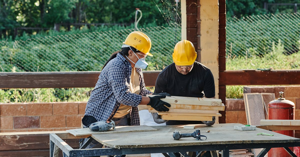Two construction workers organize wooden planks on a table at a sunny outdoor site.