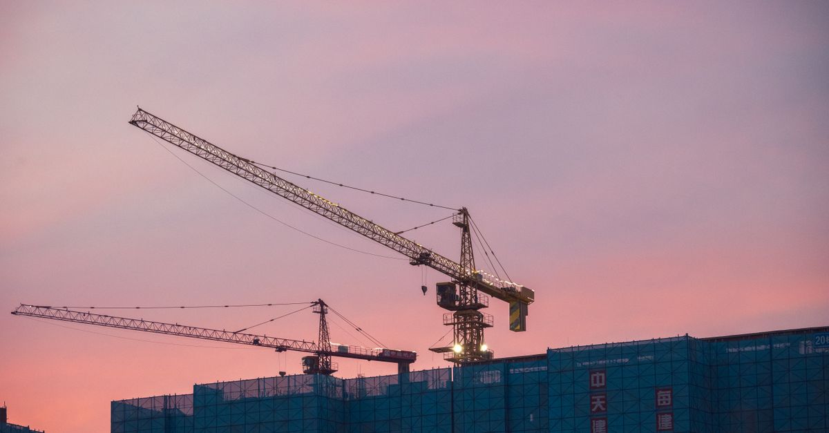 Construction cranes tower over a city skyline at sunset, symbolizing urban growth.