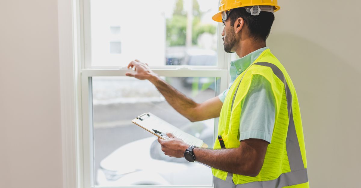 A construction worker in a safety vest and hard hat inspects a window indoors, holding a clipboard.