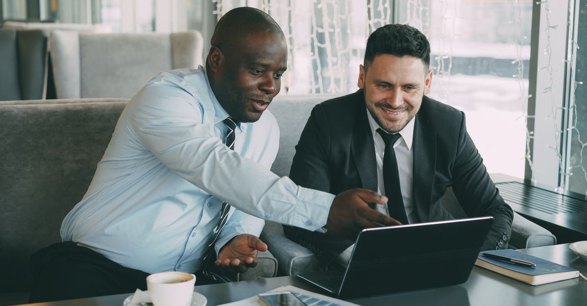 Two businessmen discussing work while using a laptop in a cafe setting.