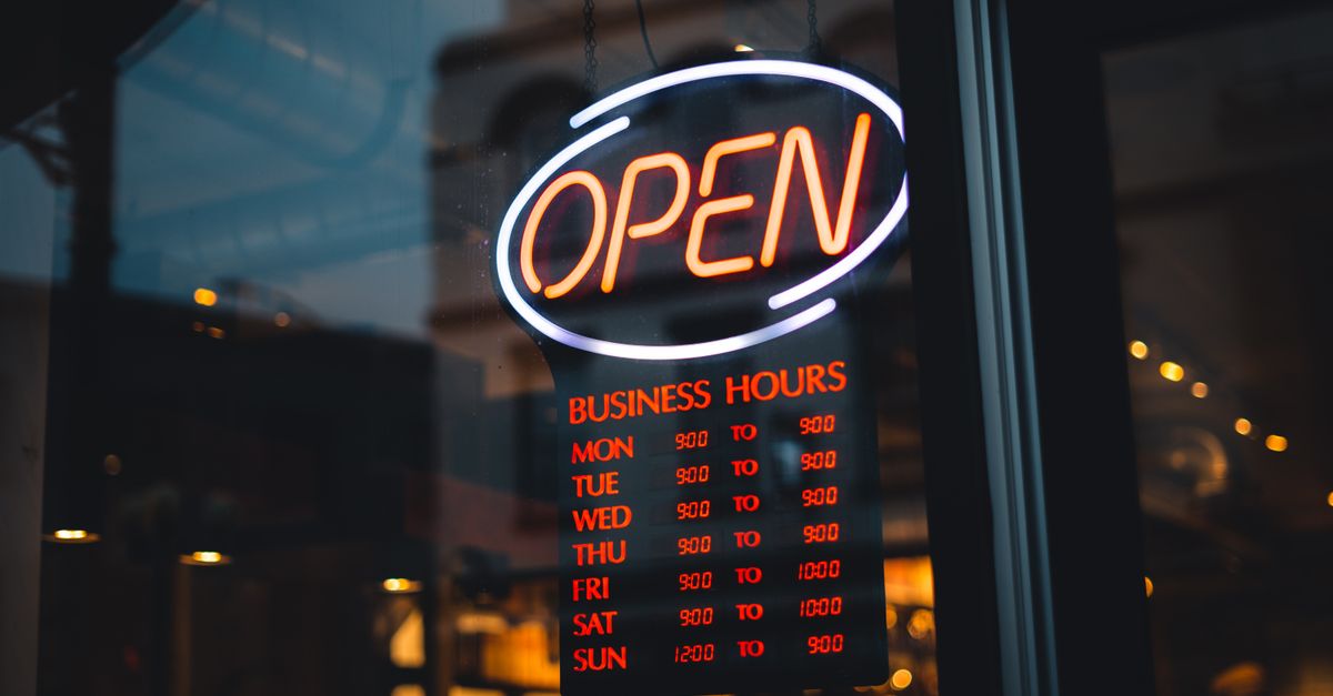 A glowing neon 'OPEN' sign with detailed business hours in a storefront window at night.