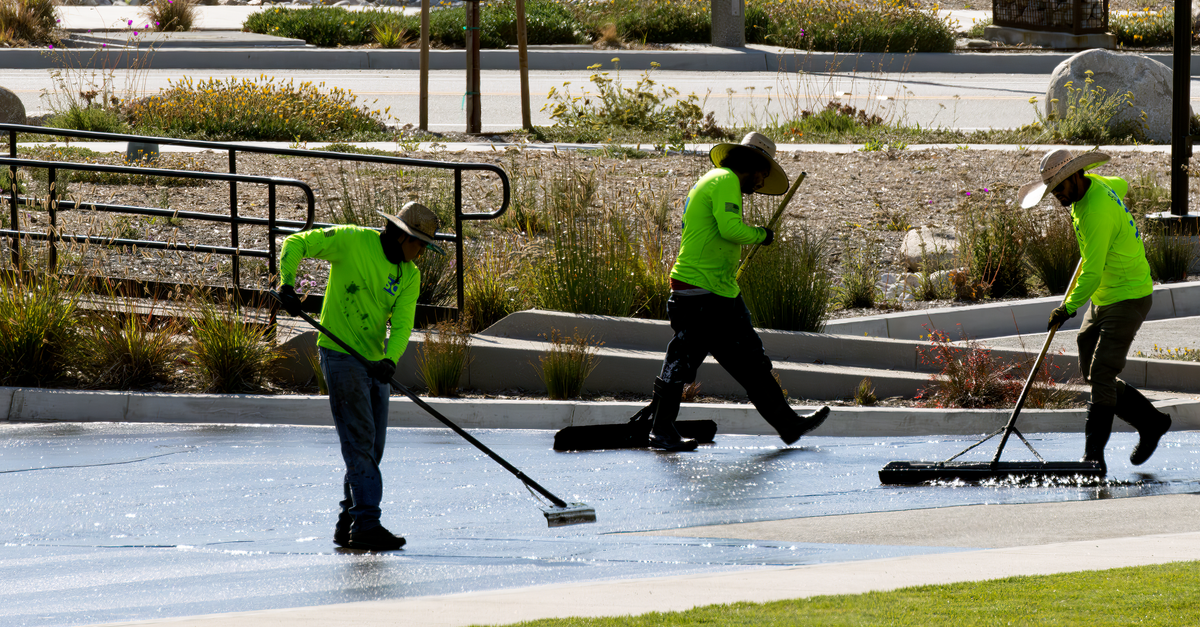Crew of workers in bright uniforms cleaning a park walkway with brooms under sunlight.