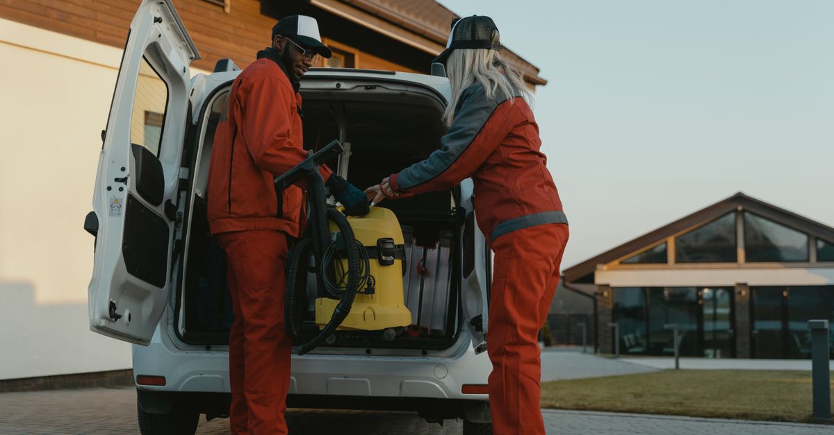 Two cleaners loading a vacuum into a van outside a modern building at dusk.