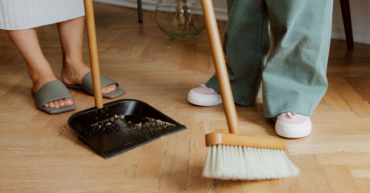 Close-up of people cleaning wooden floor with broom and dustpan indoors.