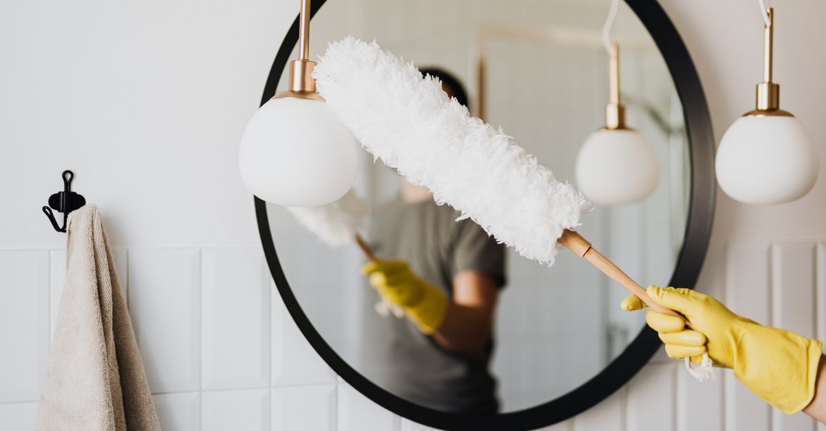 Crop anonymous female housewife in rubber gloves dusting modern lamp with soft brush against mirror while cleaning stylish bathroom
