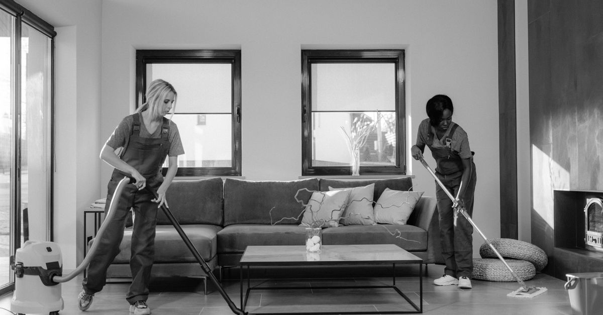 Two women in work clothes cleaning a modern living room using a vacuum and mop.