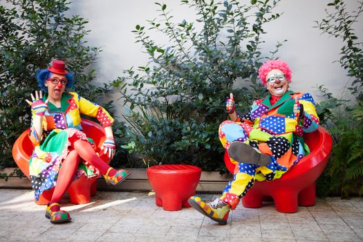 Vibrant clowns in colorful costumes sitting outside with red chairs, spreading joy and humor.