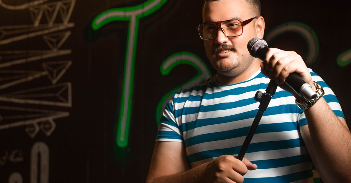 Man holding a microphone doing stand-up comedy in casual striped shirt on stage.