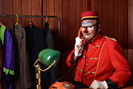 A clown in a red costume and makeup using an orange vintage rotary telephone indoors.