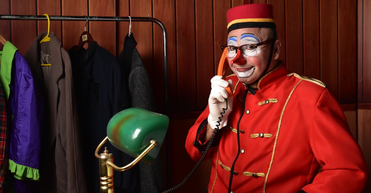 A clown in a red costume and makeup using an orange vintage rotary telephone indoors.