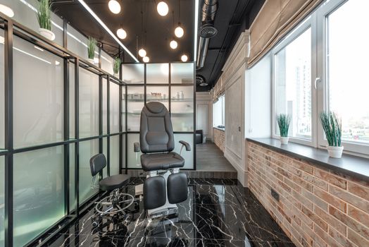 Black leather pedicure chair placed between glass door and window in modern beauty salon with decorated plants and glowing lamps