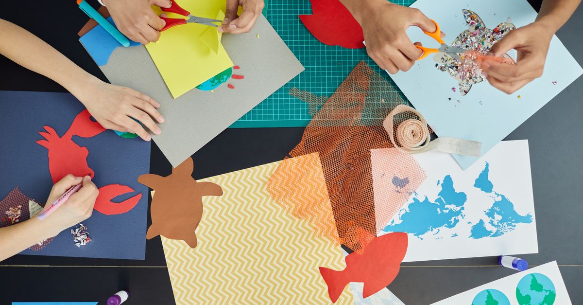 Top view of group of crop anonymous children sitting at table and creating colorful draft and artworks with paper and scissors
