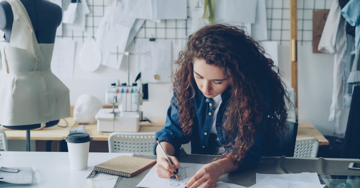 Young female fashion designer sketching in a stylish studio setting with mannequins and sewing equipment.