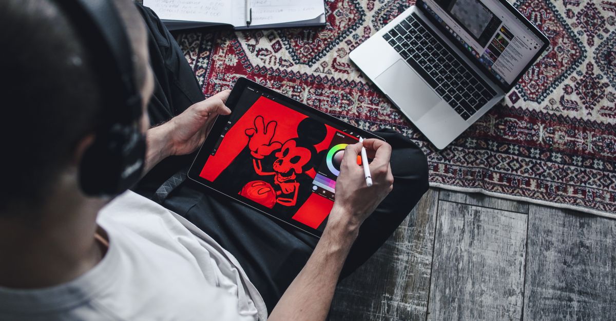 Person using a digital tablet for drawing and design work at home with a laptop.