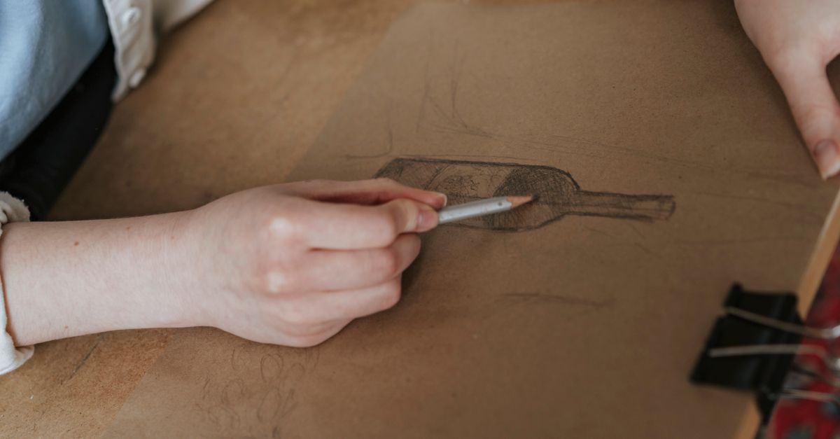 A person sketching a wine bottle on paper with a pencil. Artistic process in a creative setting.