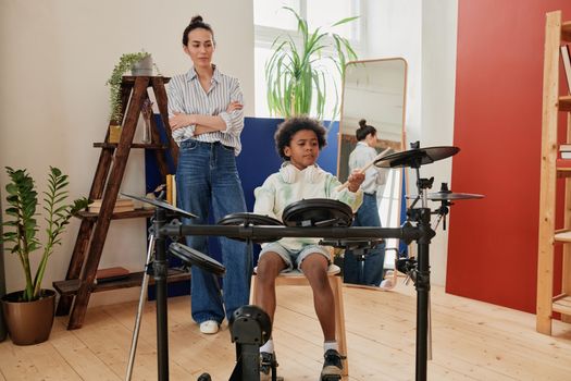 A young boy plays electronic drums under the supervision of a woman in a cozy indoor setting.