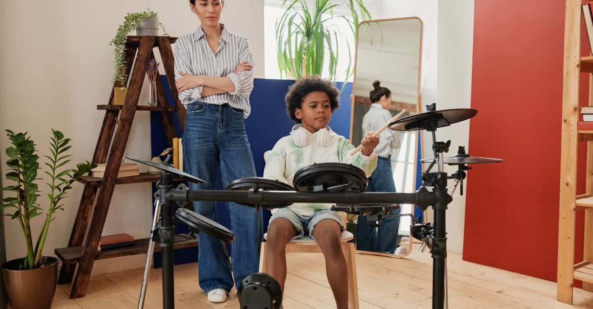 A young boy plays electronic drums under the supervision of a woman in a cozy indoor setting.