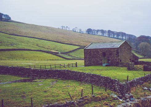 A solitary stone barn on a grassy hillside with surrounding dry stone walls on a cloudy day.