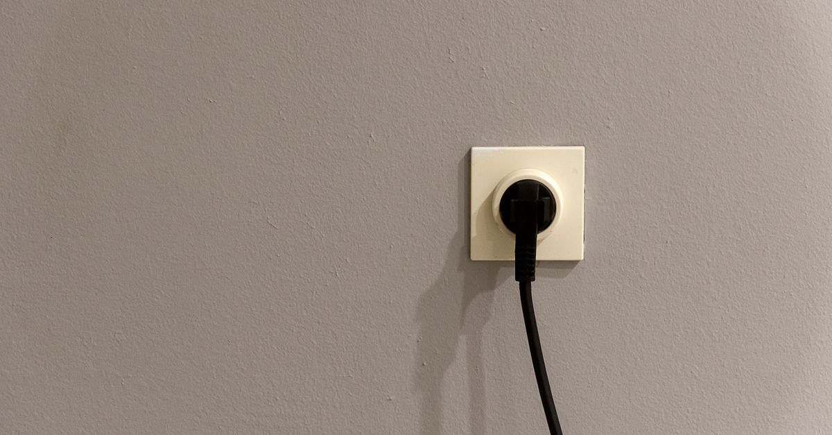 A black plug inserted into a white wall socket on a plain grey wall.
