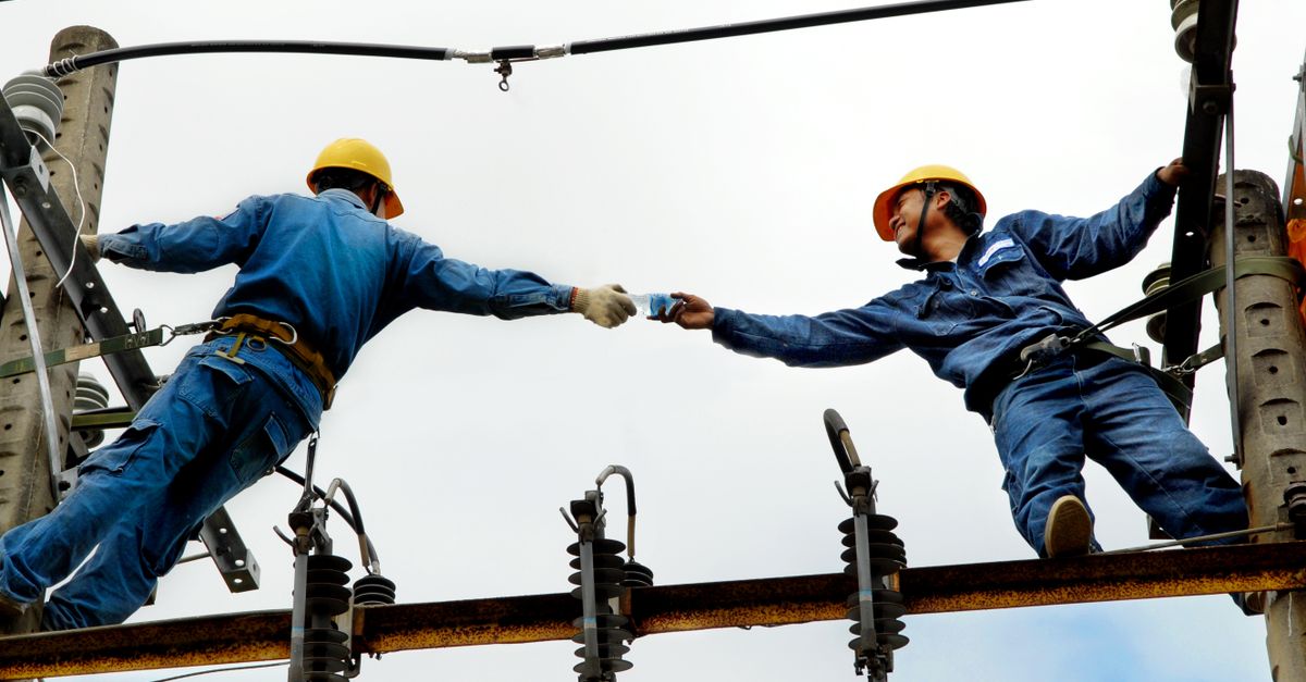 Two electrical workers in blue uniforms collaborating on power lines during daytime.