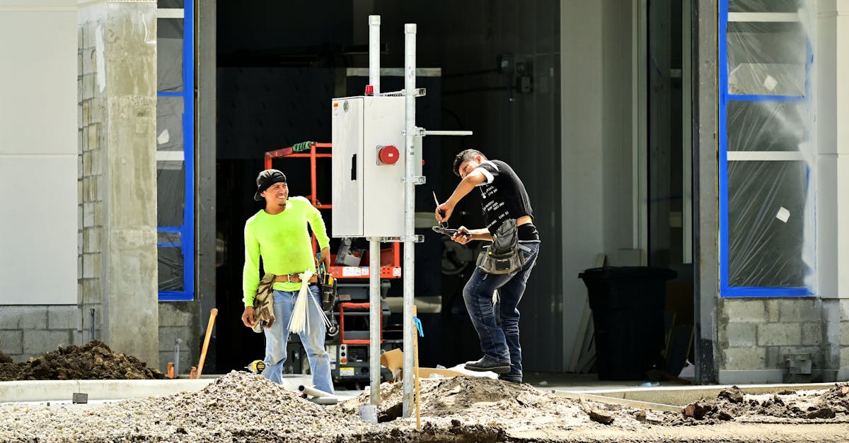 Two construction workers install an electrical panel outside a building site, wearing safety gear.