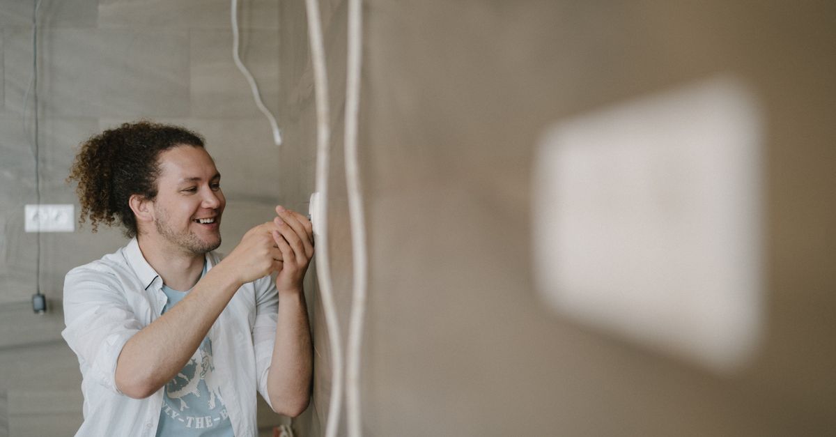An electrician with curly hair adjusts an outlet indoors, smiling as he works.