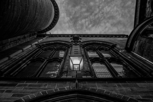 A striking black and white photograph of a gothic building with arched windows and a central lamp, set against a textured sky.