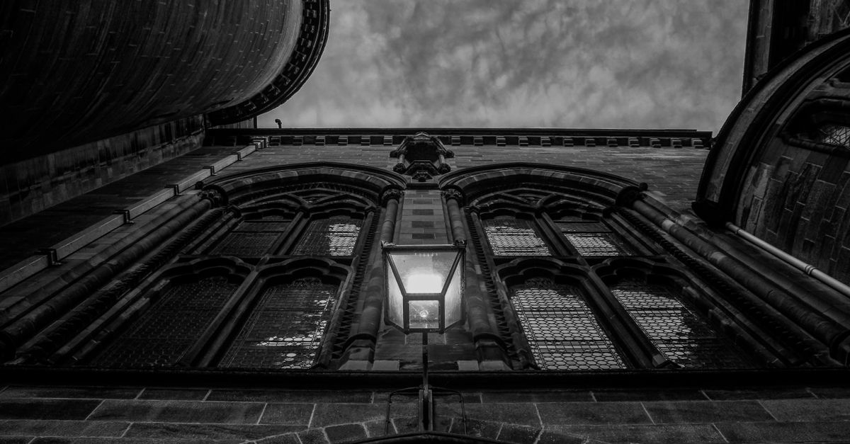 A striking black and white photograph of a gothic building with arched windows and a central lamp, set against a textured sky.