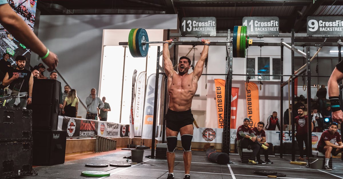 A muscular athlete performs a powerful overhead lift during a CrossFit competition indoors, showcasing strength and determination.