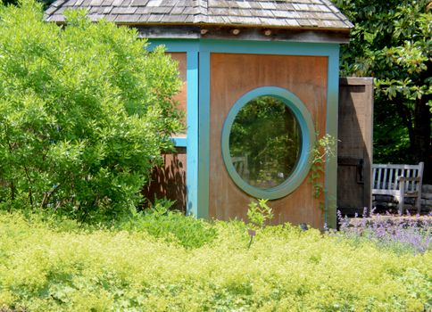 Charming garden hut with circular window surrounded by lush greenery on a sunny day.