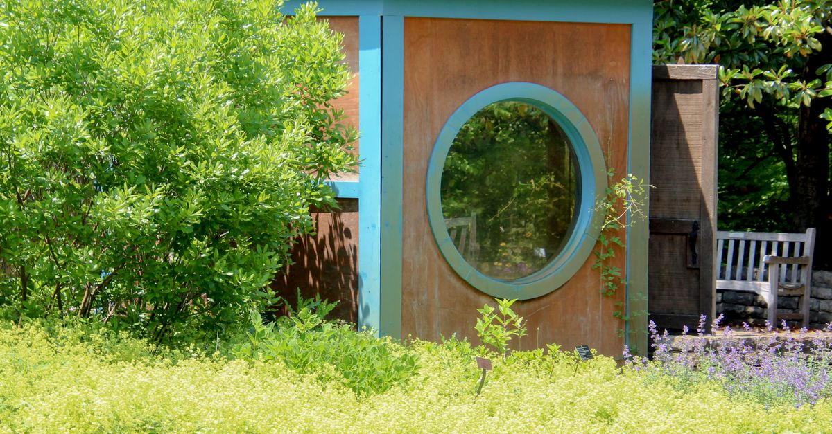 Charming garden hut with circular window surrounded by lush greenery on a sunny day.