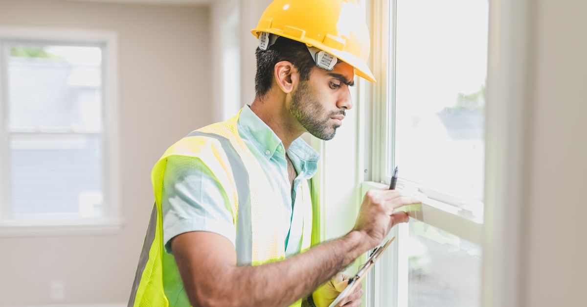 Construction worker with hard hat and vest inspecting window indoors, side view.