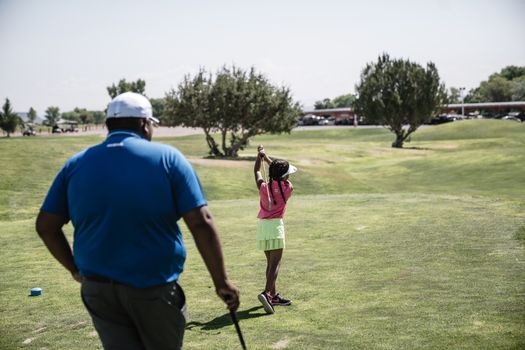 A young girl practices her golf swing under the guidance of her father on a sunny day at the golf course.