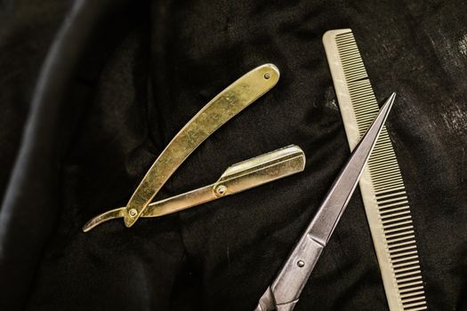 Close-up of silver shears, comb, and straight razor on a black cloth background, highlighting barber tools.