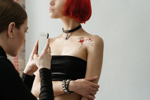 A woman with red hair and a shoulder tattoo being photographed by another woman in a tattoo studio.