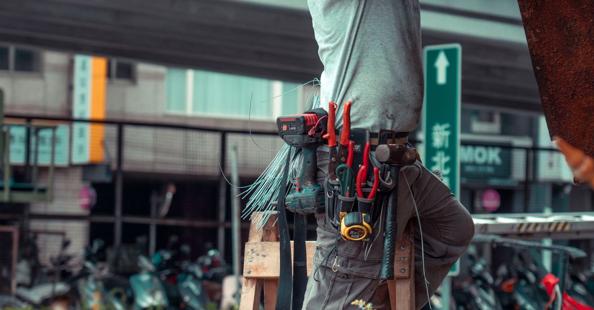 Handyman standing on a ladder with tool belt, repairing outdoors with visible tools.