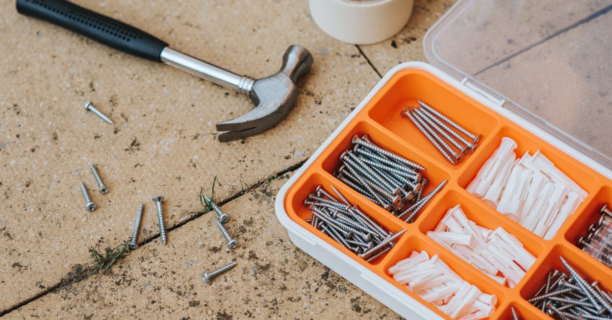 Top view of assorted repair instruments and screws in box near hammer and scotch tape on dirty tile in soft daylight
