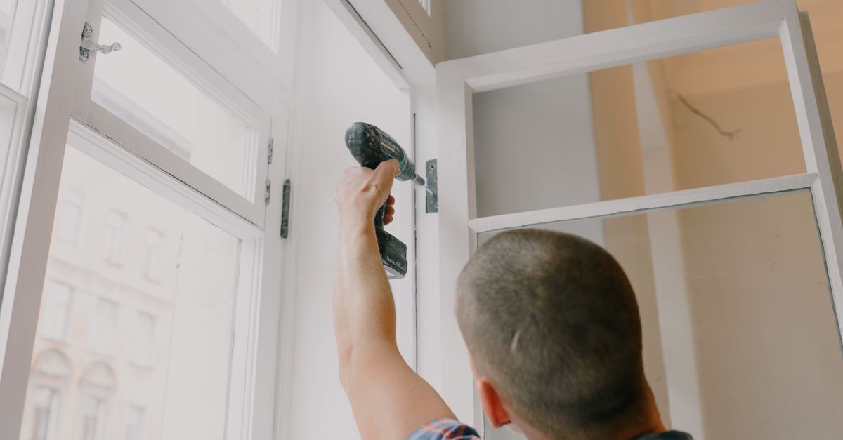 Back view of unrecognizable craftsman using cordless screwdriver and assembling window while screwing metal hinge during renovation works in apartment