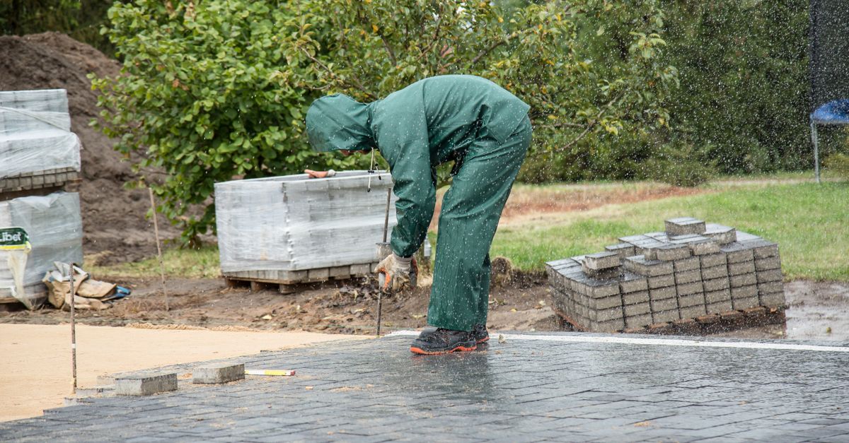 A worker in protective gear lays paving stones on a wet day.